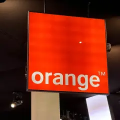 Jasny, pomarańczowy znak z napisem "orange" wisi pod sufitem. To logo Orange, które często pojawia się na ekranach telewizorów.