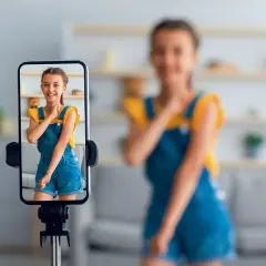 Dziewczynka na ekranie telefonu uśmiecha się, podczas gdy mama w tle nagrywa filmik. To przykład zjawiska sharenting co to znaczy dzielenie się życiem dzieci w sieci.