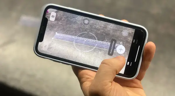 Dłoń trzyma telefon z włączoną aplikacją miarka. Na ekranie widać wirtualną linijkę i opcję dodania prostokąta.