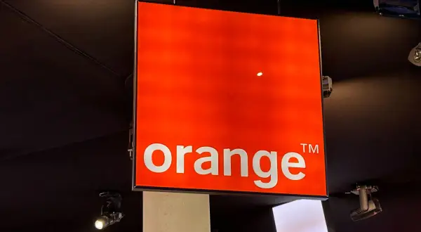 Jasny, pomarańczowy znak z napisem "orange" wisi pod sufitem. To logo Orange, które często pojawia się na ekranach telewizorów.
