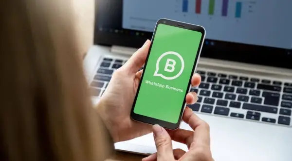 Dłoń trzyma smartfon z zielonym ekranem i logo WhatsApp Business. W tle widać laptop z wykresem.