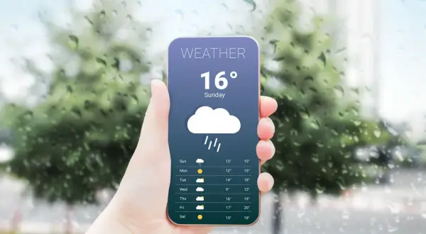 Dłoń trzyma smartfon z aplikacją pogodową pokazującą deszcz i 16 stopni. Za oknem widać krople deszczu.