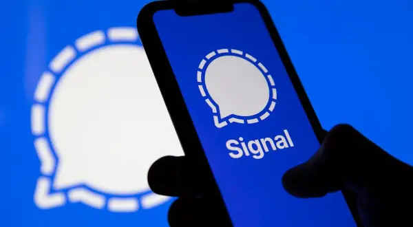 Dłoń trzyma smartfon z otwartą aplikacją Signal. W tle widoczne logo Signal.