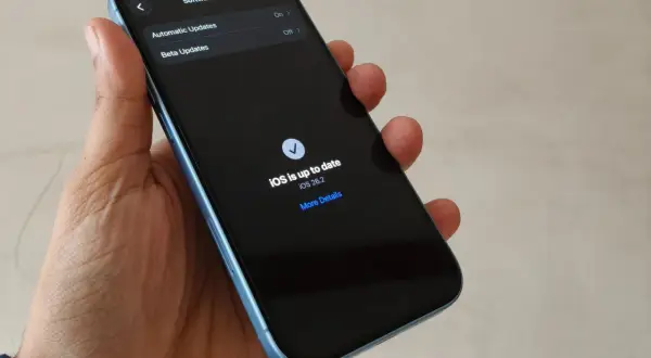Dłoń trzyma iPhone'a z ekranem pokazującym "iOS is up to date iOS 26.2".