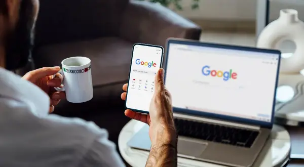 Mężczyzna z kubkiem kawy przegląda instrukcję, jak wylogować się z konta Google, na telefonie i laptopie.