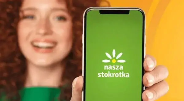 Uśmiechnięta kobieta prezentuje ekran telefonu z zieloną aplikacją "Nasza Stokrotka".