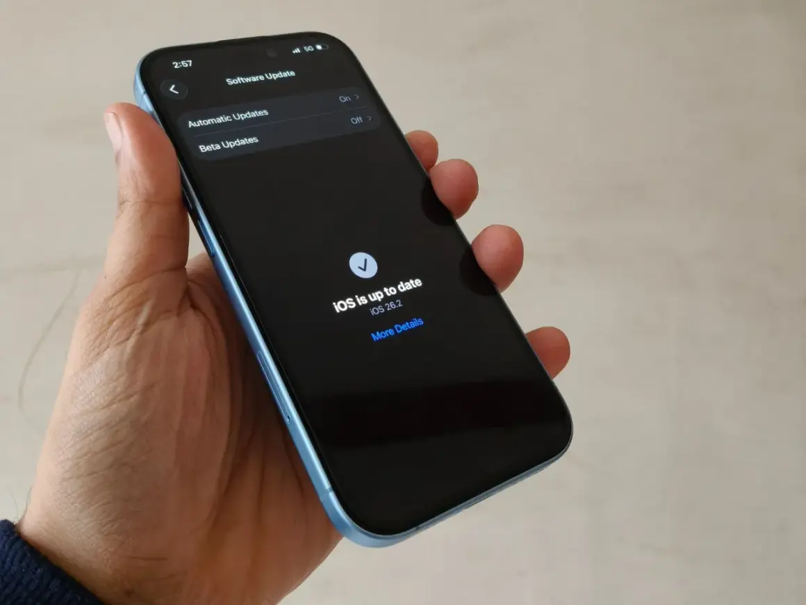 Dłoń trzyma iPhone'a z ekranem pokazującym "iOS is up to date iOS 26.2".