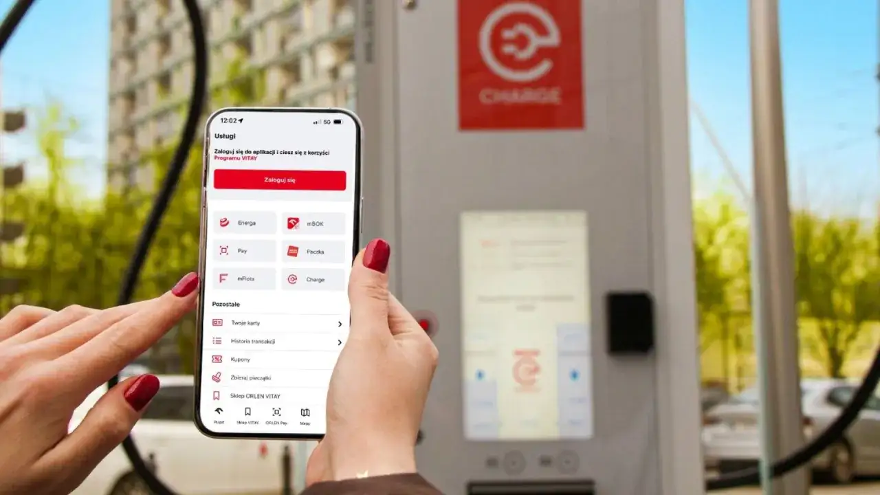 Kobieta używa aplikacji Energa na telefonie, by zalogować się do programu VITAY. W tle widać ładowarkę do samochodów elektrycznych.